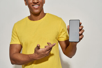 Cropped shot of smiling young man in casual yellow t shirt showing and pointing at smartphone with blank screen while standing isolated over gray background