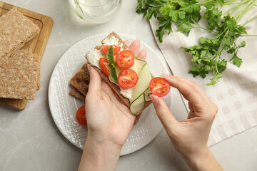 Woman eating fresh rye crispbread with cream cheese and vegetables at light grey marble table, top...