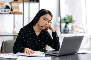 Obraz premium Upset young beautiful asian woman in glasses and headset, call center worker, manager or freelancer, sitting at table in the office, looking sadly at the laptop screen, resting her head on her hand