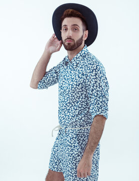 Chico joven de barba anteojos de sol y gorro posa parado con mono con im&aacute;genes estampadas 