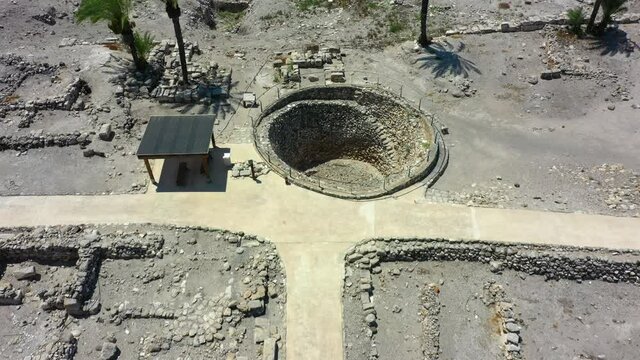 Aerial Tilt Up Shot Of Tel Megiddo Ruins With Palm Trees, Drone Flying Backward Over Protected Site Against Mountains On Sunny Day