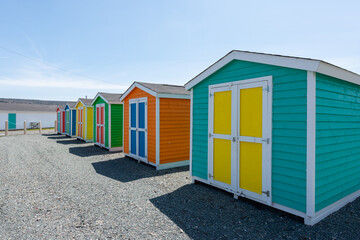 Multiple wooden sheds with colorful double doors. The buildings are blue, yellow and green storage buildings. The colorful huts have white trim on the edges. The background is a blue sky with clouds. 