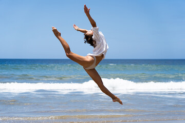 Young joyful female dancer in swimsuit and white shirt while happily jumping with sea on background. Carefree and happiness concept 