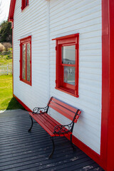 The exterior of a vintage white wooden building covered in narrow cape cod clapboard with double hung windows painted in red trim. There's an iron and wood bench on the grey deck with a green garden.