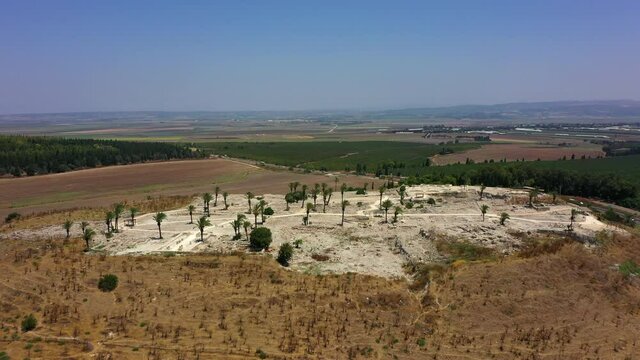 Aerial Panning Shot Of Tel Megiddo Ruins On Hill Against Blue Sky, Drone Flying Backward From Protected Site By Road On Sunny Day