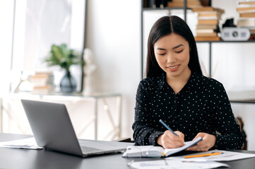 Online work with a laptop or distance learning. Young adult pretty female asian freelancer, manager or student, sitting at work desk, using laptop for work or study, taking notes during online webinar