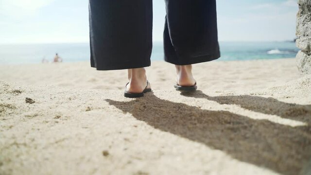Young woman in wide black pants and a hat walks along the beach to the water. Sunny day at sea. Close-up of feet in black flip flops. Warm holidays on the golden beach. Summer chill. White sand.