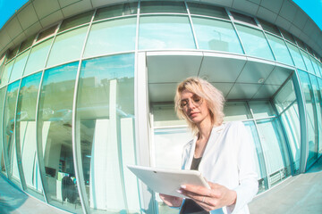 a blonde woman in a business white suit and round glasses stands with a white tablet in her hands against the backdrop of a glass building. horizontal photo. Half-length portrait. fish eye
