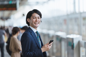 駅のホームでスマートフォンを持つ若い男性・通勤とIoTのイメージ
