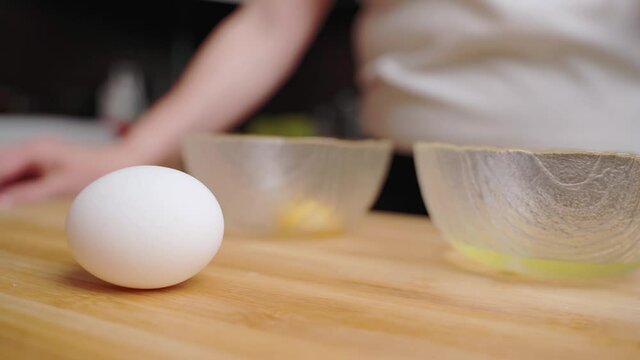 Hands of young asian woman chef cracking eggs into ceramic bowl separating egg yolk from white on wooden board at kitchen table at home. Healthy food lifestyle and traditional bakery concept.