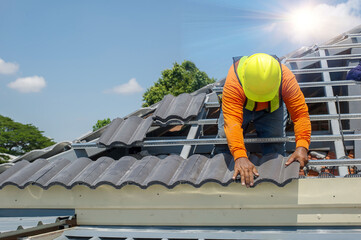 Roof repair, worker with white gloves replacing gray tiles or shingles on house with blue sky as background and copy space, Roofing - construction worker standing on a roof covering it with tiles.