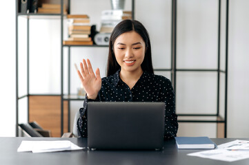 Friendly cute confident young asian woman freelancer, manager, ceo, sitting in office at laptop, talking by video call with the client or employees, discussing business strategy, waving hand, smiling