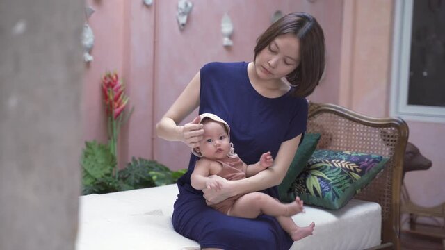 asian woman mother wearing hat for baby infant at tropical room. short hair mom adjusting baby hat with love and care.