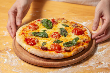 Chef holds wooden tray or board with homemade cheese pizza with tomato cherry, sauce, sausage and basil. Tasty dinner, close up view