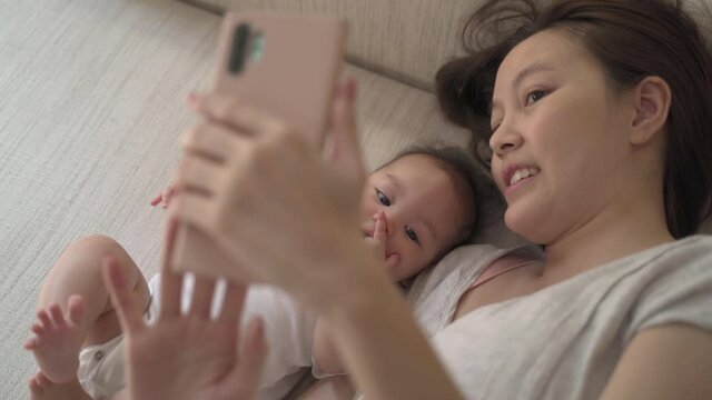 asian woman mother parent using mobile smart phone taking selfie photo with baby infant on bed at home. family video call via smartphone.  