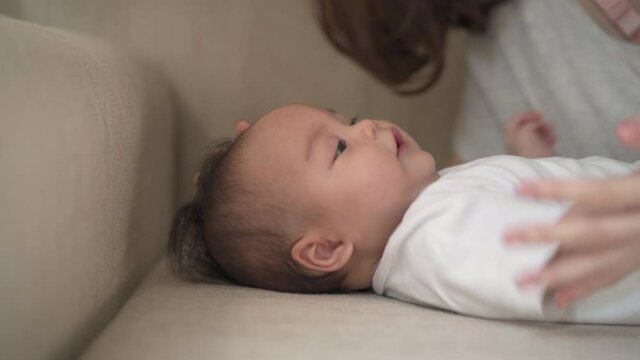asian woman mother kissing baby infant on cheek softly on bed. baby happy when mother parent kissing with love and care. mom using nose touching kiss daughter at home. authentic lifestyle.