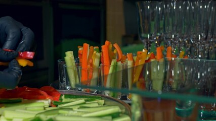 The cook puts various vegetables in glassware - yellow, red bell peppers and fresh cucumbers cut into strips, the work is done in black nitrile gloves.