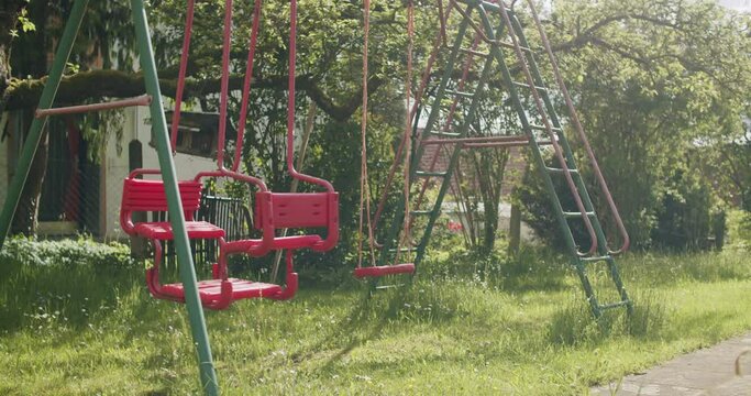 Wide shot of a typical German swing set in the garden on a sunny spring day. Swing moving in the wind, nobody in the frame. Static shot with cinematic backlight from the sun.
