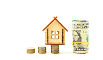 Miniature toy wooden house stands on top of a stack of metal coins, US dollars rolled into a tube, isolated on a white background. Concept of buying and selling apartments, houses. Copy space.