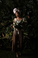 Fototapeta premium Charming calm curly hair, dark - skinned woman in white dress posing in a tropical decoration, holding a bouquet of colorful flowers in her hands