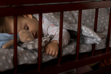 Cute cheerful little chubby baby girl sleeping sweetly in grey baby crib during lunch rest time in white and pink pajamas with teddy bear at home. Childhood, leisure, comfort, medicine, health concept