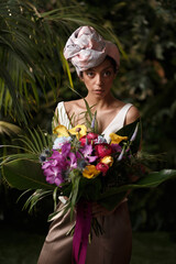 Charming calm curly hair, dark - skinned woman in white dress posing in a tropical decoration, holding a bouquet of colorful flowers in her hands