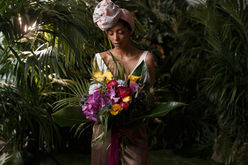 Charming calm curly hair, dark - skinned woman in white dress posing in a tropical decoration, holding a bouquet of colorful flowers in her hands