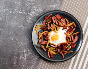 Fried potatoes with sausage and egg on a round plate on a dark background. Top view, flat lay.
