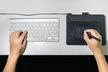 A young man is working on a computer on a desk using a keyboard and a pen to work.