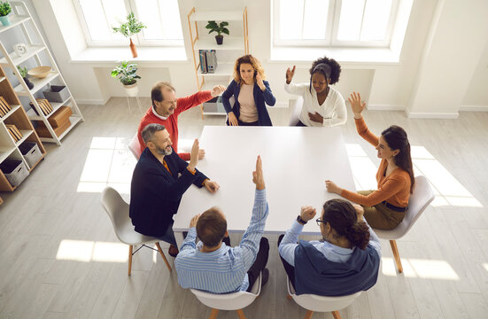Team of happy united people raising hands and voting. Group of young and mature colleagues unanimously accepting good suggestion in corporate meeting around office table. From above, high angle
