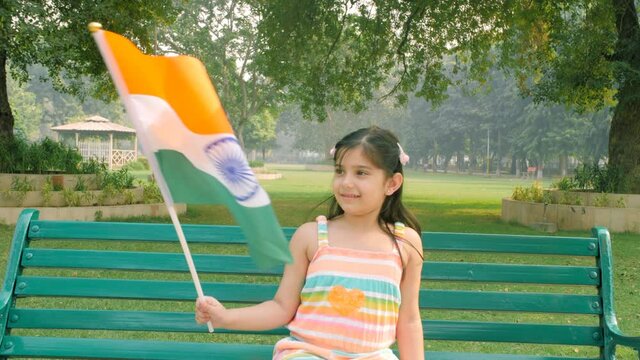 School going Indian girl waving the flag of India - Independence day / Republic day. Little cute girl celebrating Independence day by waving the Indian tricolor sitting on the bench in the park - p...