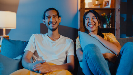 Romantic asia couple man and woman smile and laugh lay down on sofa in living room at night watch...
