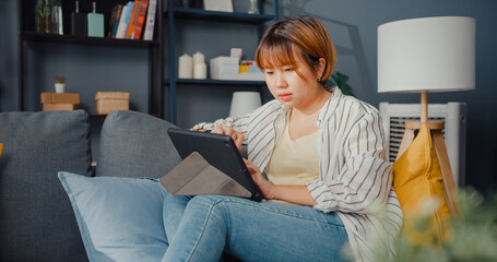 Freelance Asia lady casual wear using tablet online learning in living room at house. Working from home, remotely work, distance education, social distancing, quarantine for corona virus prevention.