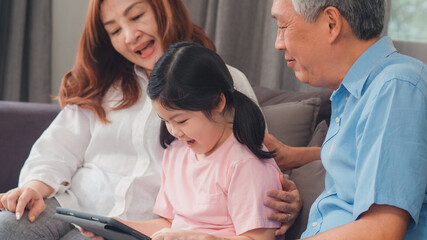 Asian grandparents and granddaughter using tablet at home. Senior Chinese, grandpa and grandma happy spend family time relax with young girl checking social media, lying on sofa in living room concept