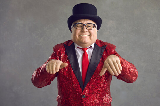 Smiling Rich Senior Man In The Mood To Party. Funny Positive Old Fat Millionaire In Glasses, Bling Fancy Sequin Suit And Black Top Hat Looking At Camera, Dancing And Vibing To Music On Gray Background