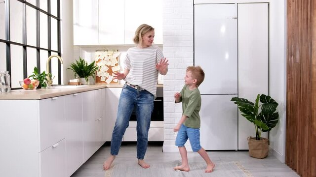 Excited Mom Or Babysiter And Kid Boy Jumping, Dancing, Laughing In Modern Scandinavian House Kitchen. Happy Family Mother And Son Having Fun Enjoy Playing Singing Together At Home