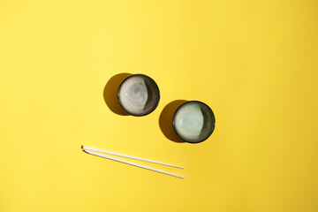 Two small grey bowls for sushi sauces and two chopsticks on yellow surface