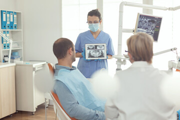 Obraz na płótnie Canvas Orthodontist nurse holding digital tablet with tooth radiography on screen explaining to sick man stomatology treatment to prevent toothache. Patient sitting on dental chair in modern dentistry office