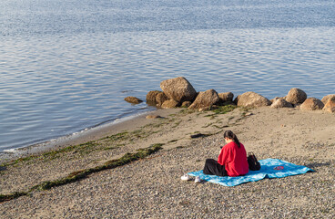 Back view of the young women in the red sweatshirt sitting on the blanket by the beach.