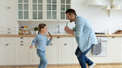 Overjoyed young Caucasian father have fun sing in kitchen appliances with small daughter. Happy dad...