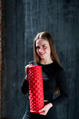 Portrait of a girl Pilates trainer with a red massage roll in her hands, stands on a gray background.