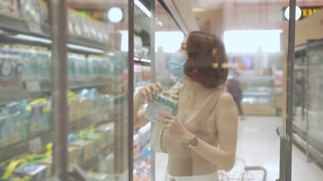 asian woman in medical face mask shopping in supermarket in covid-19 pandemic. female choosing products in modern grocery store. woman decision buying nutrition fresh food. milk and yogurt in fridge. 