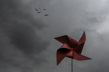 A red pinwheel against a dramatic sky with dark clouds and a flock of birds flying high in the sky