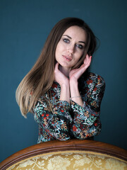 A brunette stands behind a golden chair in a mysterious floral dress