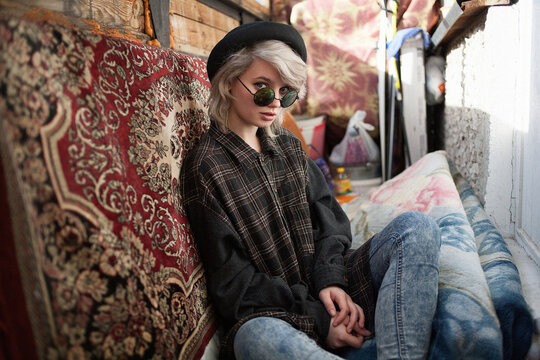 young attractive caucasian female with short hair in checkered shirt, blue jeans and black hat sitting at old vintage balcony among mess, surrounded by different junk