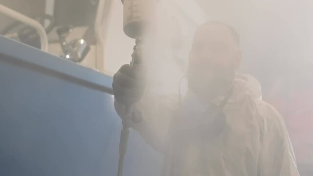 Chest-up of bearded man wearing white hazmat suit and respirator hanging on neck, looking carefully, then starting using airbrush, spraying paint on camera