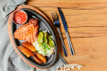 Juicy grilled pork steak with baked potatoes, cut pickles and tomato sauce served in a tray over rustic wooden table.