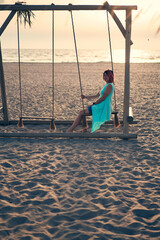 Young attractive woman with pink hair is resting on seesaw on the beach at sunset.