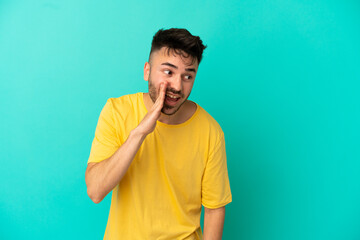 Young caucasian man isolated on blue background whispering something with surprise gesture while looking to the side