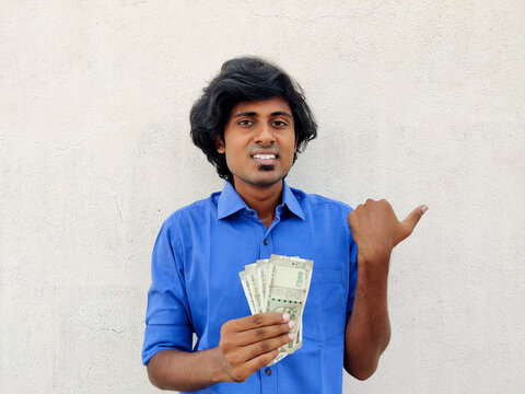 Clean shaved young handsome man wearing blue shirt holding four 500 rupees notes and pointing with thumb up to the side with happy face smiling. White background